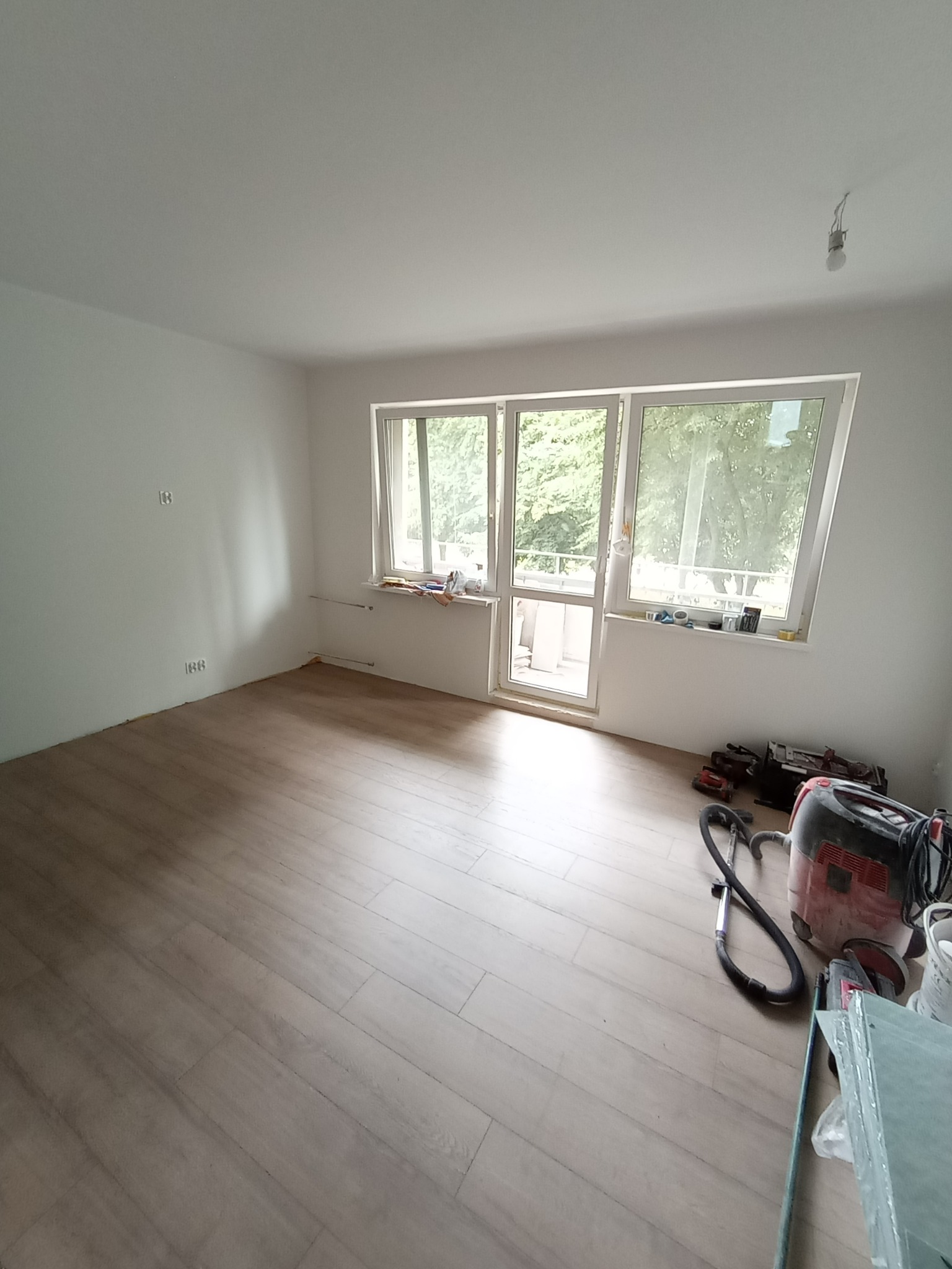 Jasne panele podłogowe w pokoju z białymi ścianami i oknem balkonowym. Widoczny odkurzacz przemysłowy i narzędzia, sugerujące prace wykończeniowe.