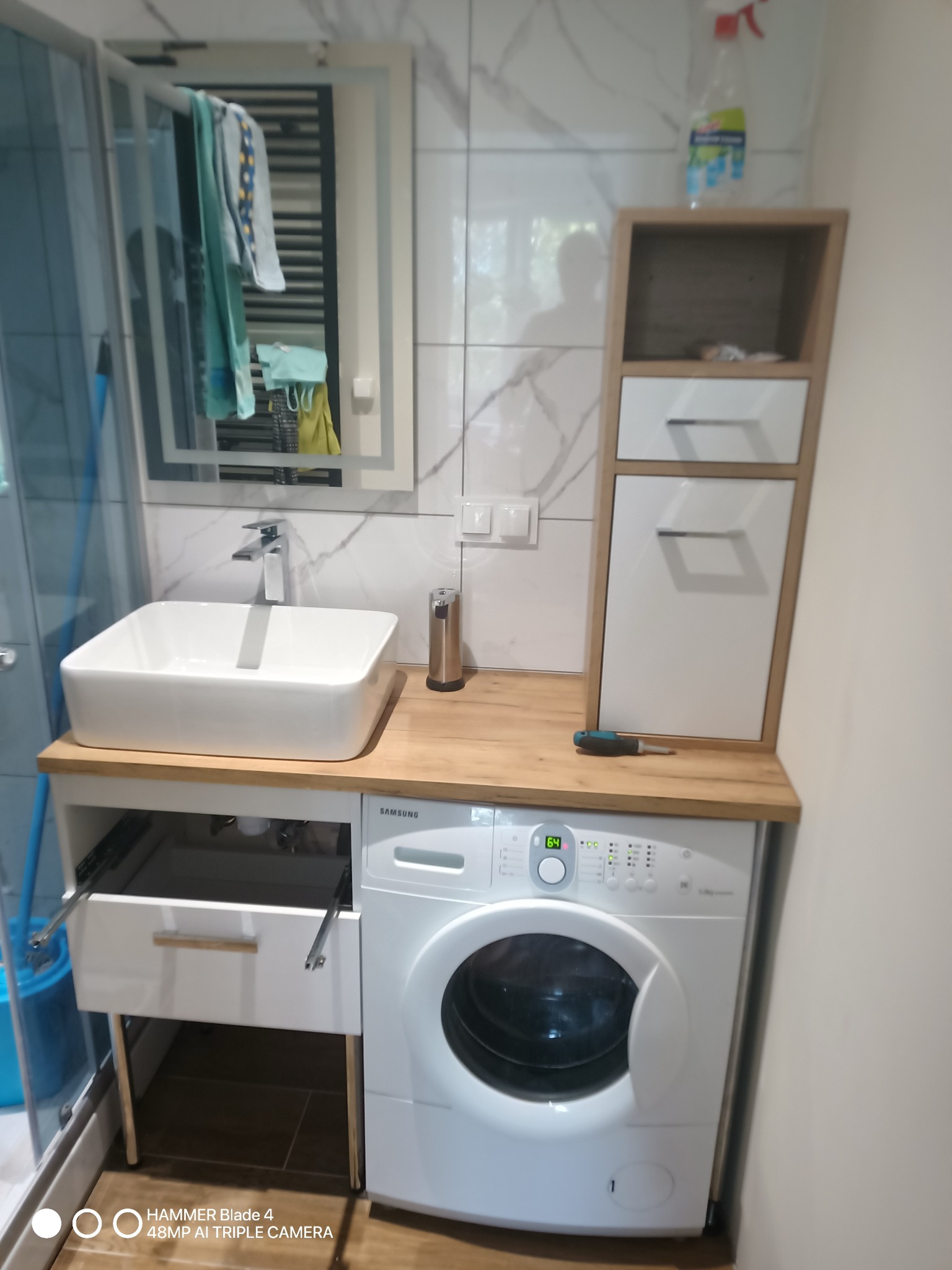 Nowoczesna łazienka z pralką zabudowaną pod blatem z umywalką, szafką wiszącą i lustrem. Biało-szara kolorystyka, widoczny otwarty szuflada i narzędzie na blacie.