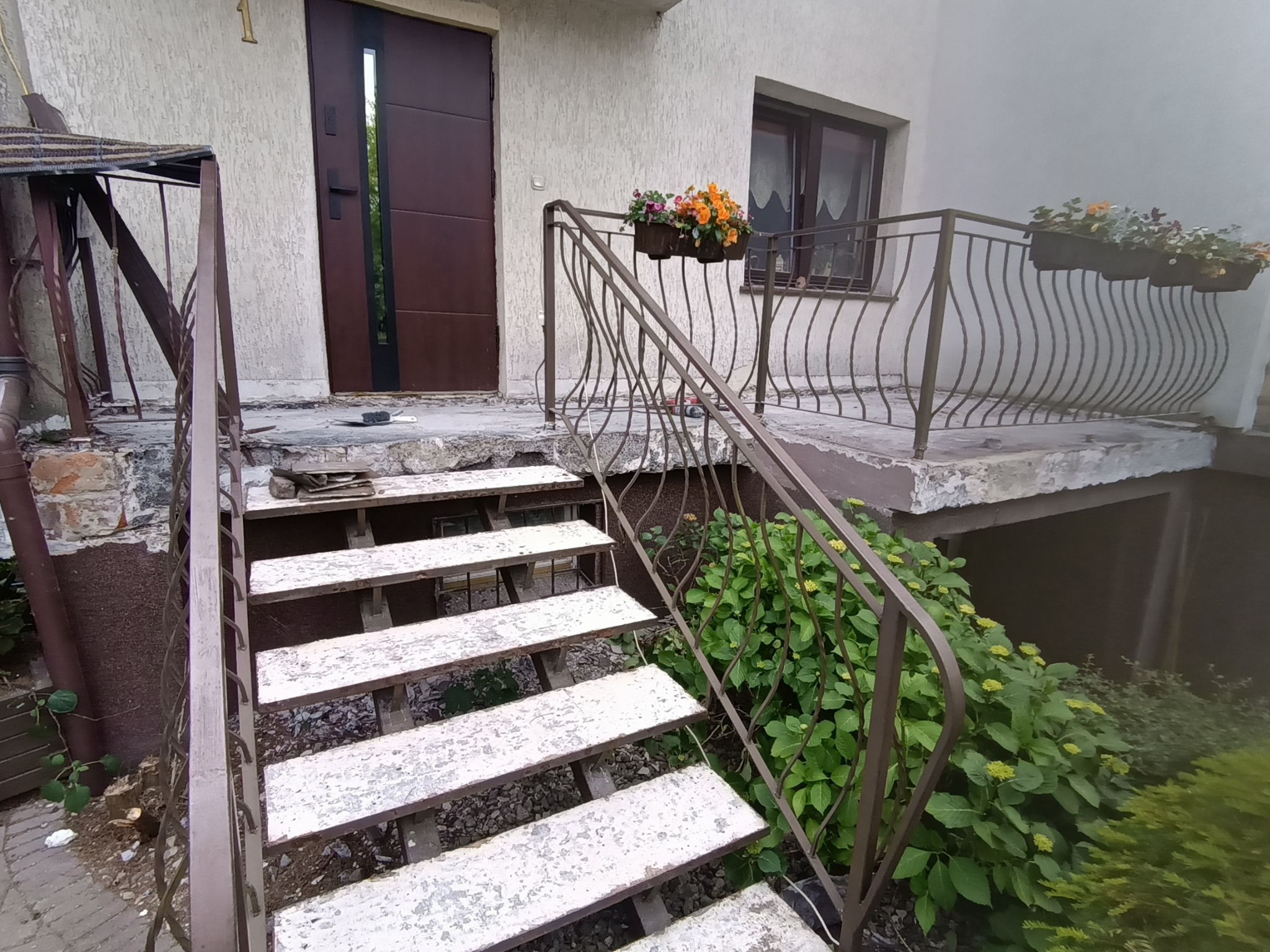 Wejście do domu z metalowymi schodami i balustradą, donice z kwiatami. Widoczne ślady użytkowania na schodach i elewacji. Numer domu '1' nad drzwiami.