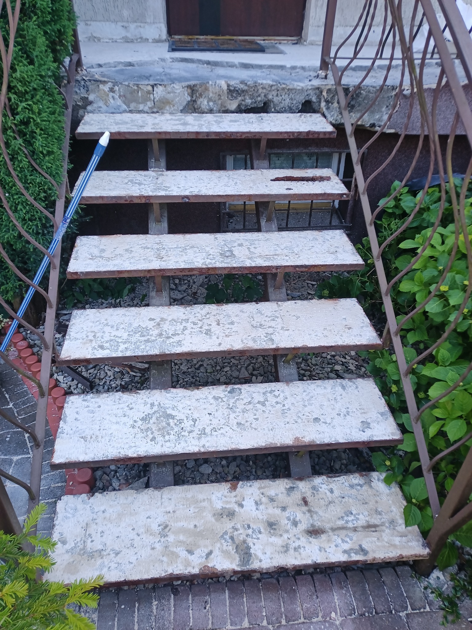 Zewnętrzne schody z metalową balustradą i betonowymi stopniami, pokryte łuszczącą się farbą. Widoczne rośliny i fragment niebieskiego przedmiotu.