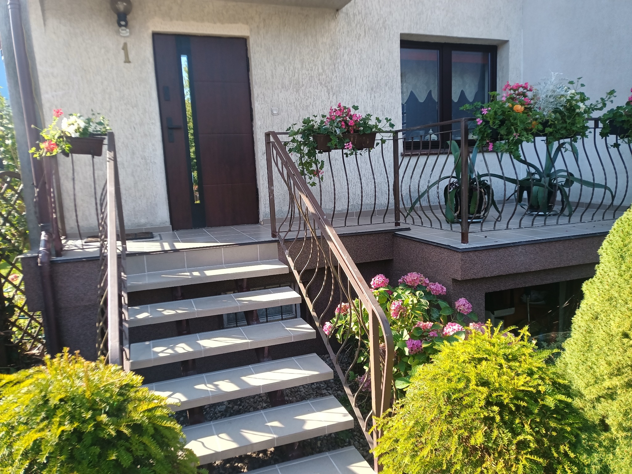 Wejście do domu z brązowymi drzwiami i schodami wyłożonymi płytkami. Balustrada z metalu, donice z kwiatami i krzewy ozdobne.
