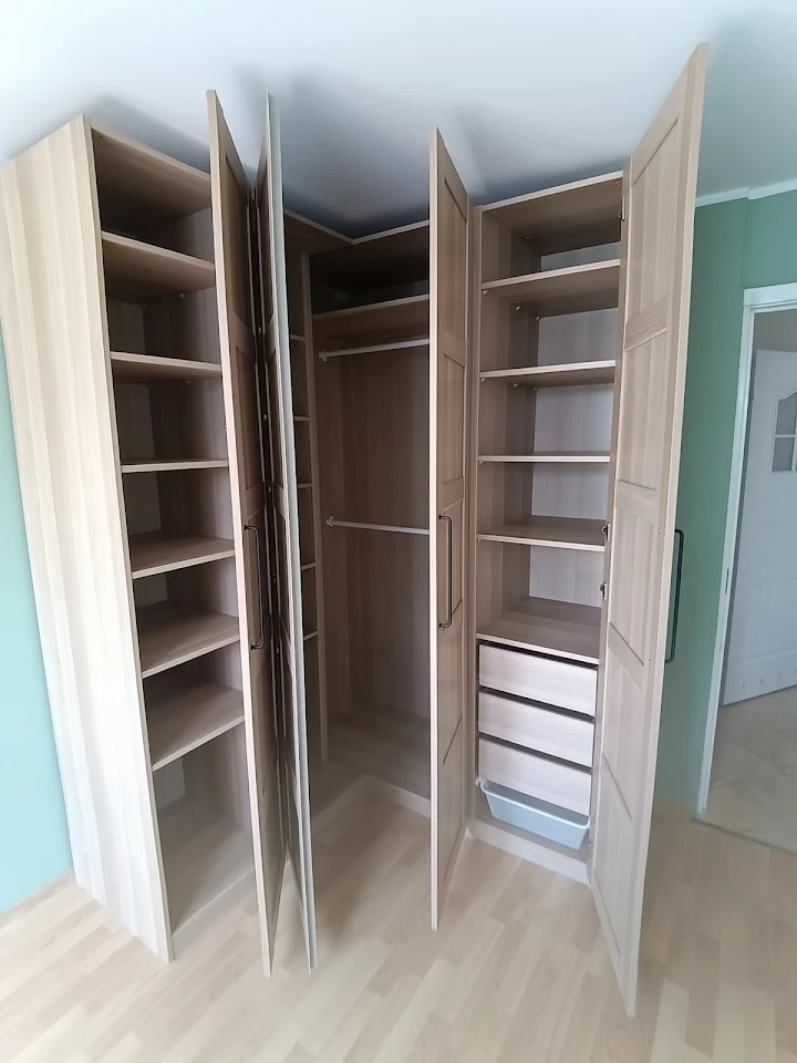 Jasny, narożny system szaf z otwartymi drzwiami, ukazujący półki, drążki i szuflady. Widoczne detale wykończenia i jasna kolorystyka drewna.