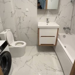 MB BUD Bohdan Mykhailiv - Biała łazienka z marmurowymi płytkami, umywalką z szafką, toaletą i pralką. W lustrze odbija się osoba robiąca zdjęcie.