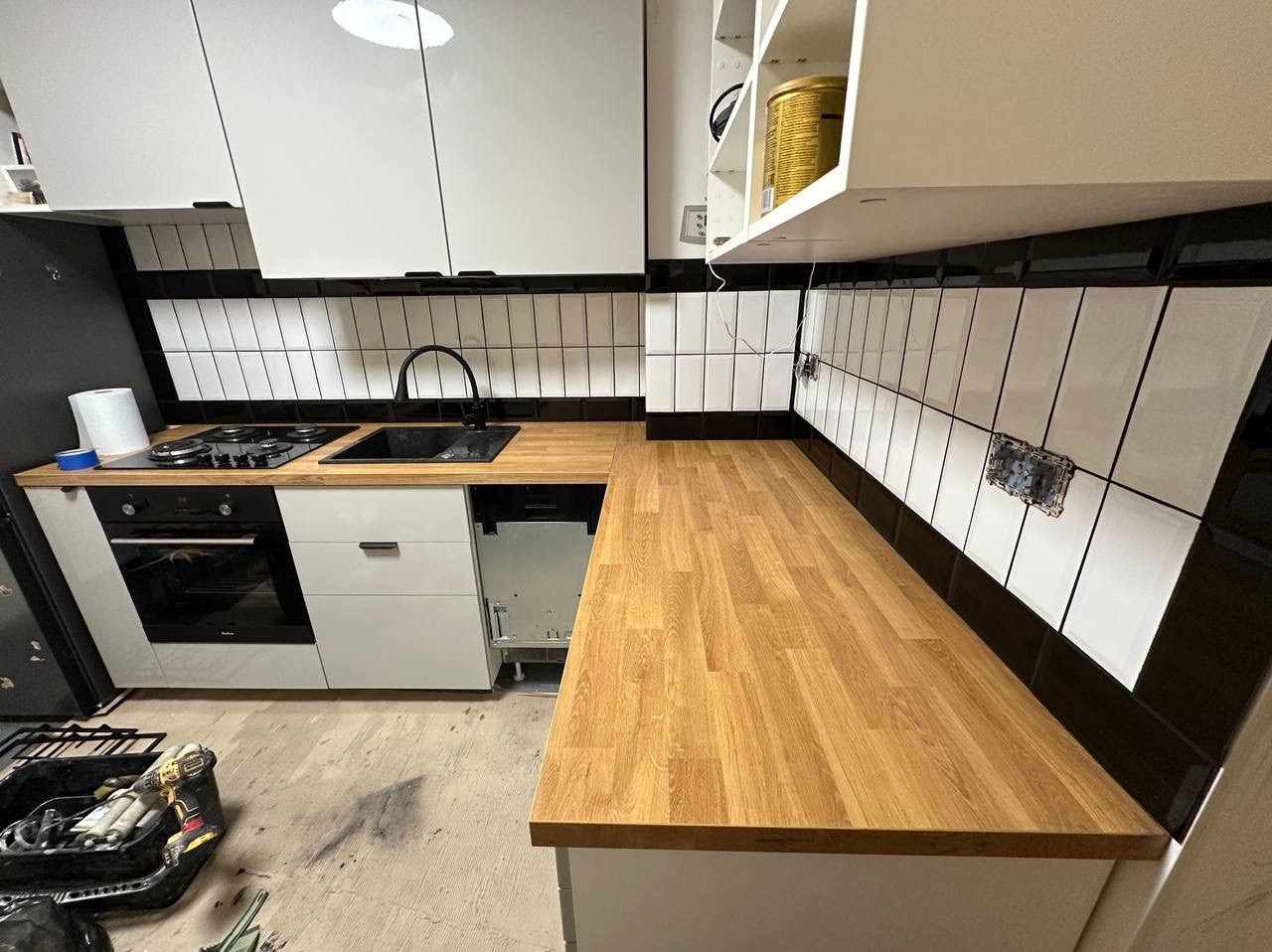 Nowoczesna kuchnia w trakcie wykończenia: białe szafki, drewniany blat, czarne akcenty. Widoczne płytki i narzędzia, sugerujące etap prac wykończeniowych.