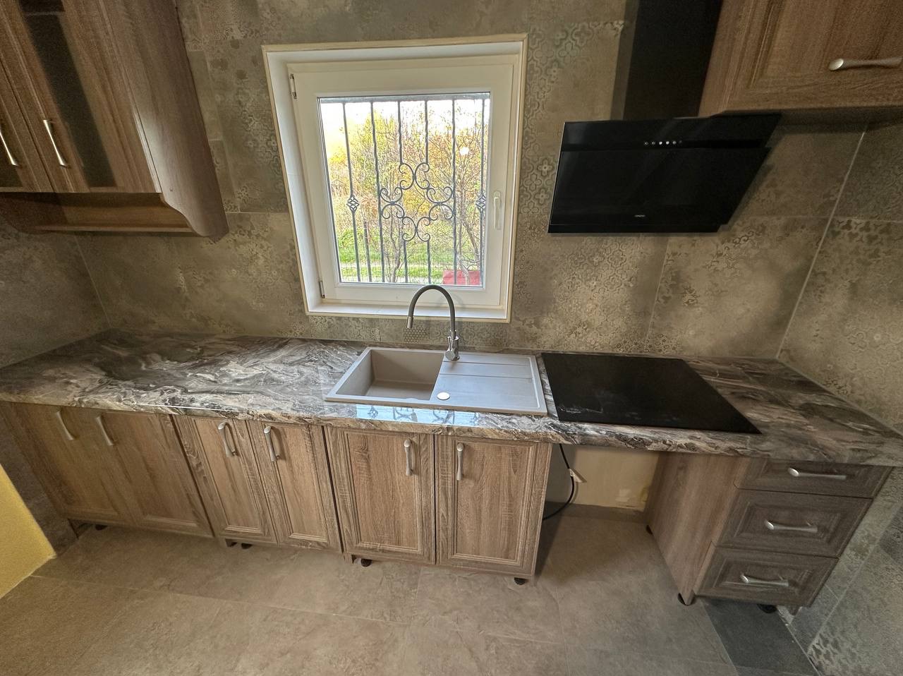 Jasna kuchnia z drewnianymi szafkami, marmurowym blatem i oknem z kratą. Nowoczesny okap nad płytą grzewczą. Płytki ścienne z delikatnym wzorem.