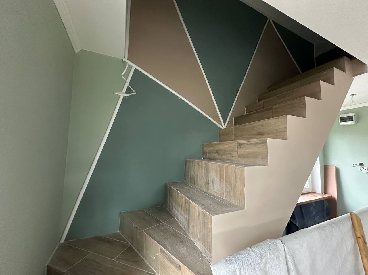 Schody wyłożone płytkami imitującymi drewno, ściana z geometrycznym wzorem w odcieniach zieleni i beżu, białe listwy dekoracyjne. Nowoczesne wykończenie.