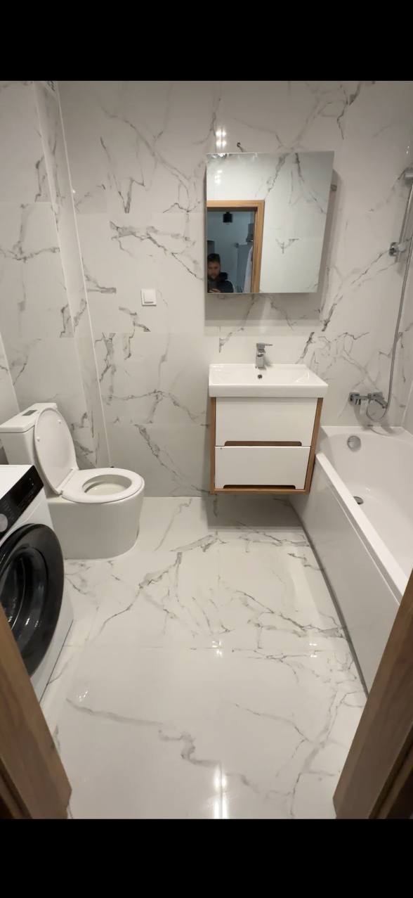 Biała łazienka z marmurowymi płytkami, umywalką z szafką, toaletą i pralką. W lustrze odbija się osoba robiąca zdjęcie.
