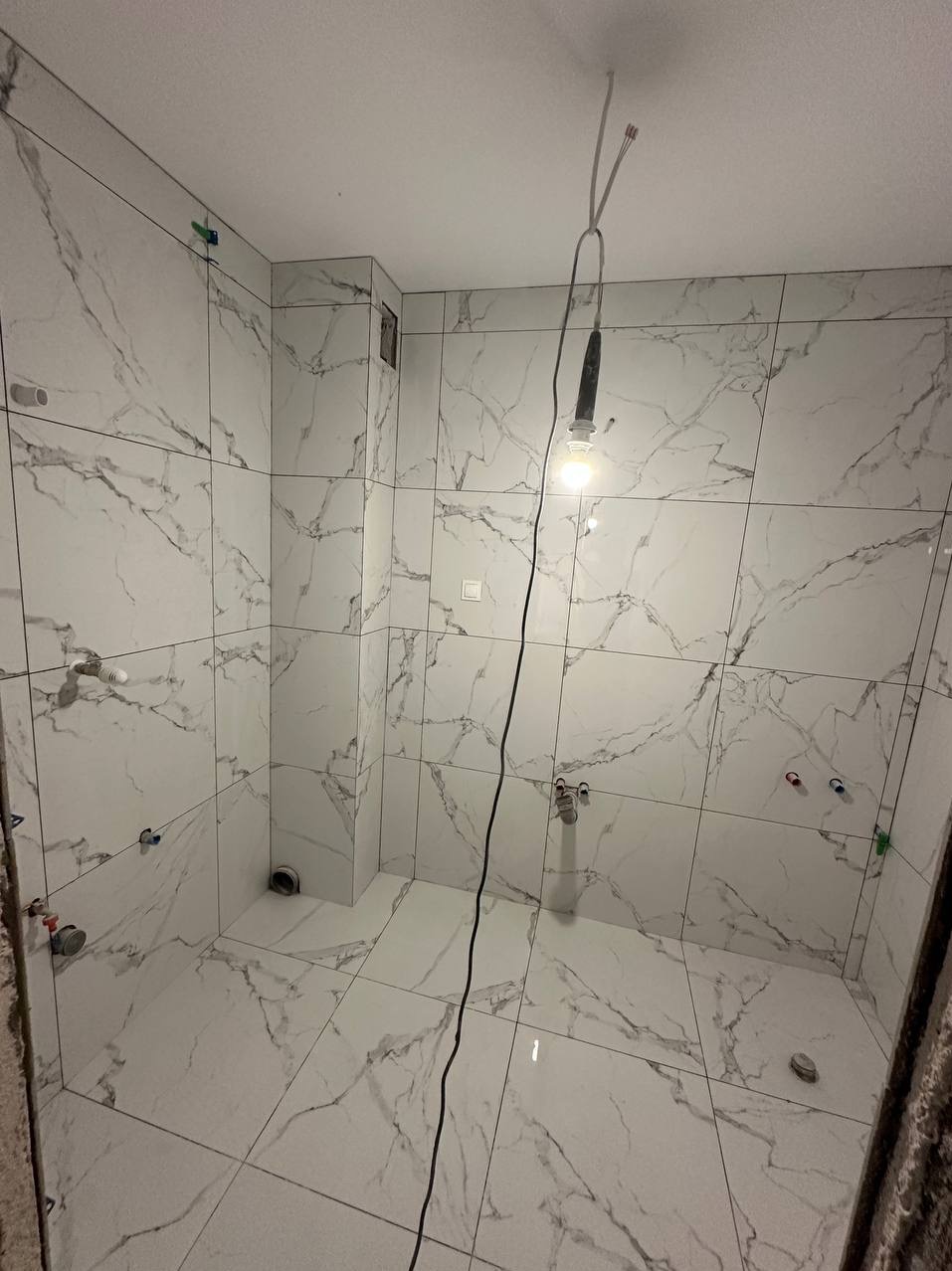 Biała łazienka z marmurowymi płytkami, widoczne przyłącza wodne i odpływy. Wnętrze w trakcie prac wykończeniowych, oświetlone wiszącą żarówką.