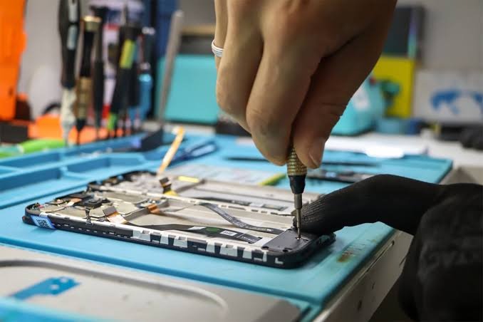 Naprawa smartfona: ręka w rękawicy precyzyjnie używa śrubokręta do rozkręcania obudowy telefonu na niebieskiej macie serwisowej, widoczne narzędzia w tle.
