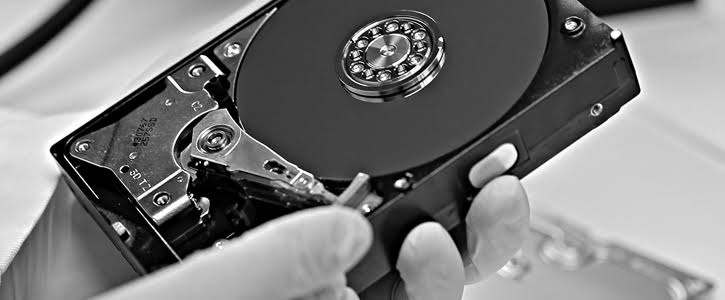 Otwarty dysk twardy w dłoniach w rękawiczkach, widoczne talerze i głowica. Czarno-biała fotografia, detale mechaniczne, precyzyjna praca.