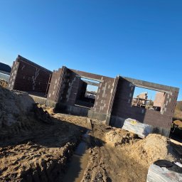 Firma Ogulno Budowlana Zbigniew Płotka - Wznoszone ściany domu z pustaków ceramicznych na fundamencie, widoczne palety z materiałem budowlanym i wykop pod instalacje. Błękitne niebo w tle.