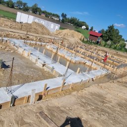 Firma Ogulno Budowlana Zbigniew Płotka - Zalane betonem fundamenty domu jednorodzinnego z drewnianym szalunkiem i wystającymi prętami zbrojeniowymi, widok z góry, w tle budynki i drzewa.