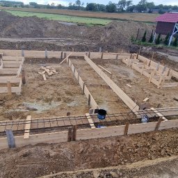 Firma Ogulno Budowlana Zbigniew Płotka - Szeroki kadr na szalunek fundamentów zbrojonych stalą, przygotowany do zalania betonem. W tle ziemia, budynek gospodarczy i zieleń.