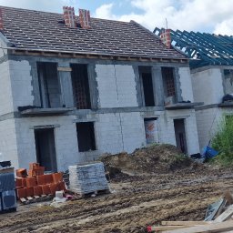 Firma Ogulno Budowlana Zbigniew Płotka - Budowa bliźniaka z białych bloczków: stan surowy otwarty, widoczne elementy więźby dachowej, palety z materiałami budowlanymi na błotnistym terenie budowy.