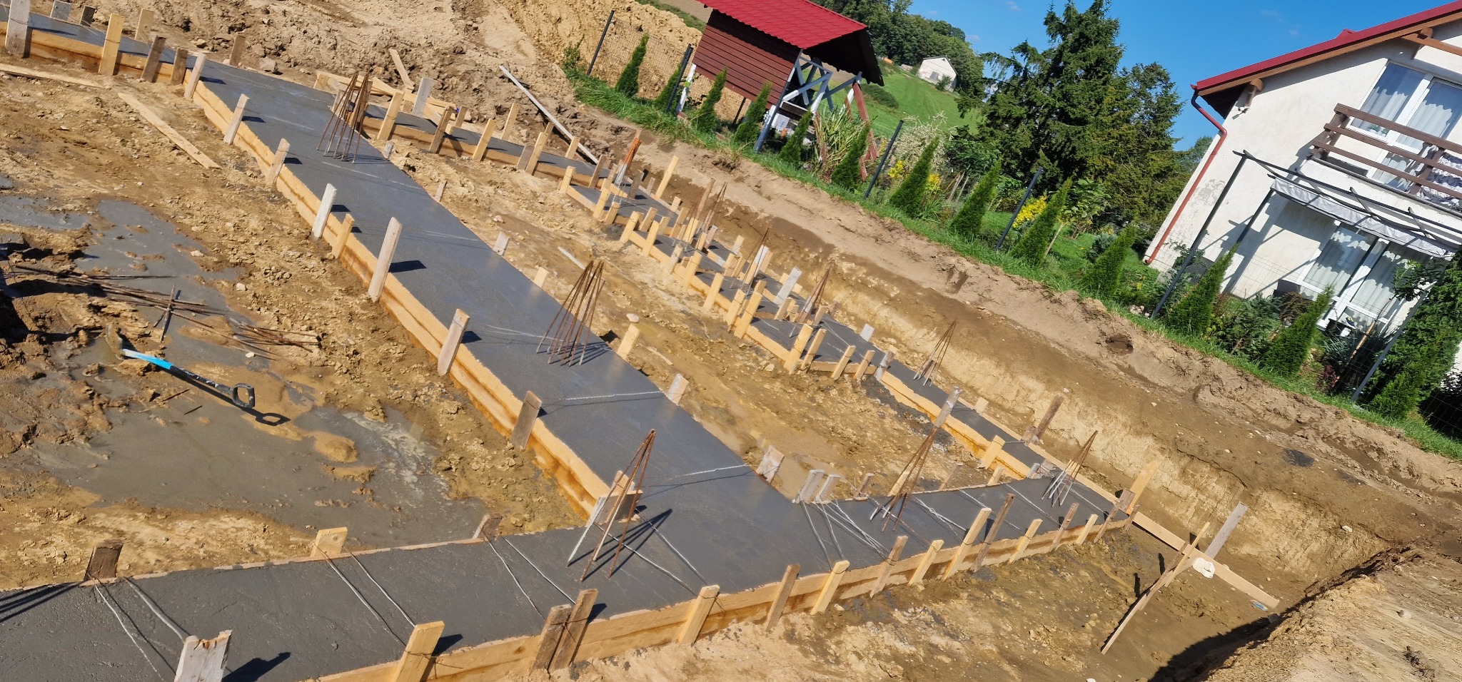 Zalane betonem fundamenty z drewnianym szalunkiem i wystającymi prętami zbrojeniowymi, widoczne z góry na tle krajobrazu z domami i zielenią.