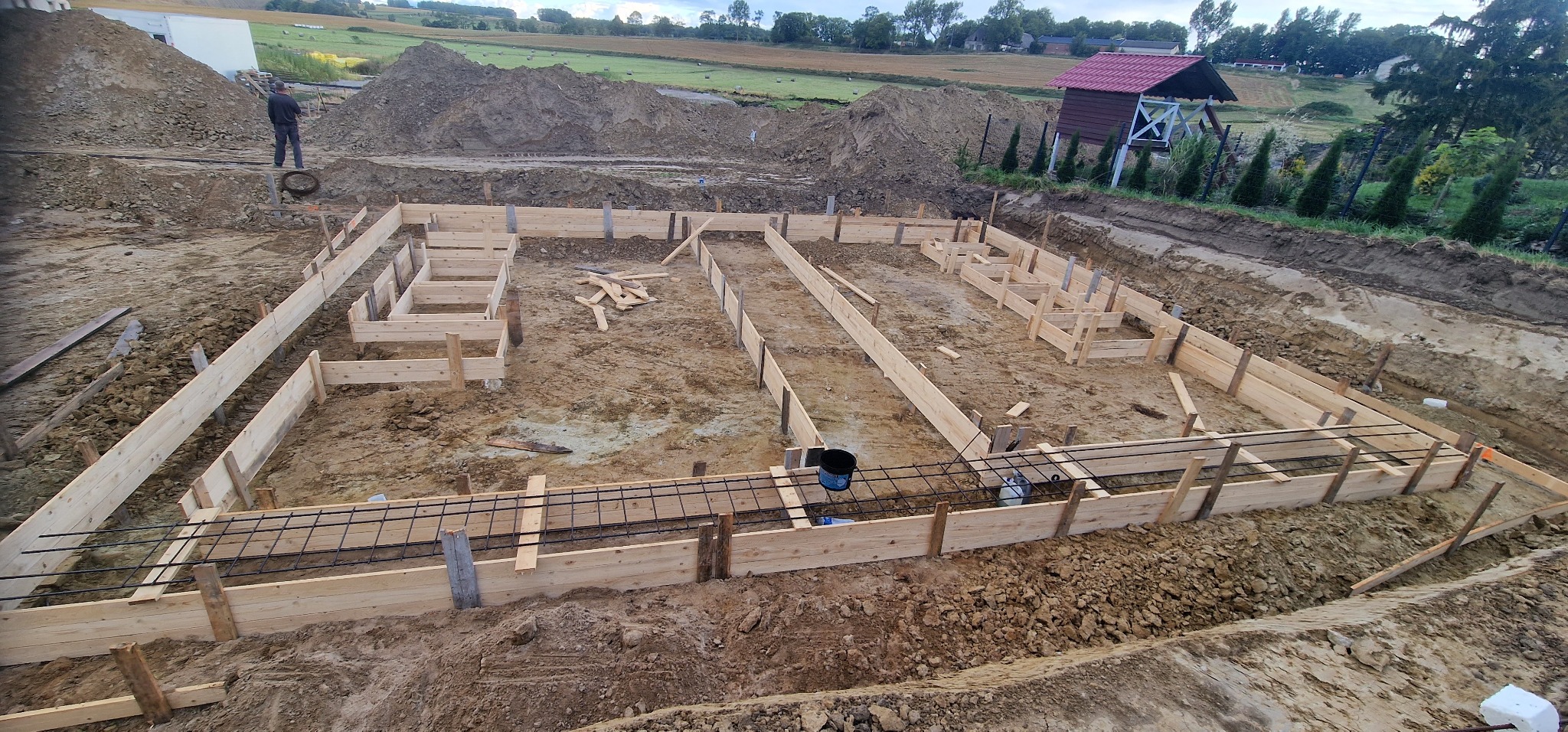 Szeroki kadr na szalunek fundamentów zbrojonych stalą, przygotowany do zalania betonem. W tle ziemia, budynek gospodarczy i zieleń.