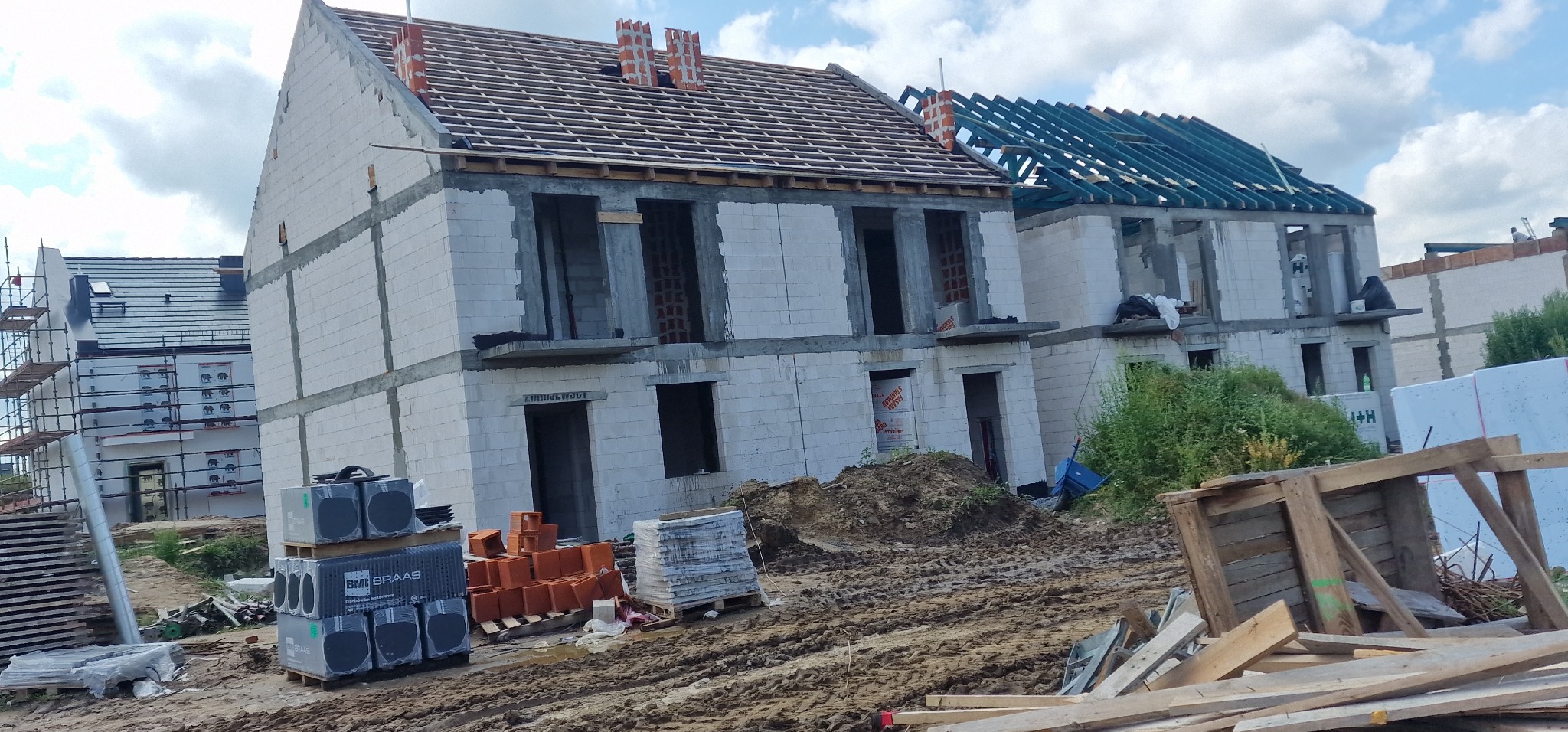 Budowa bliźniaka z białych bloczków: stan surowy otwarty, widoczne elementy więźby dachowej, palety z materiałami budowlanymi na błotnistym terenie budowy.