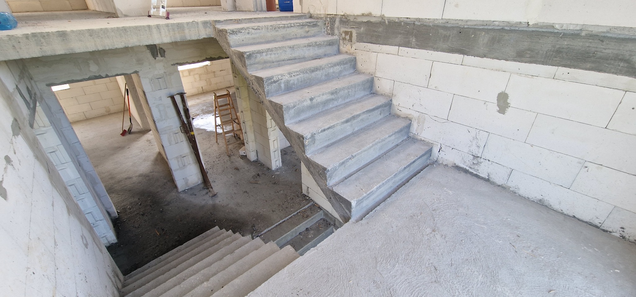 Surowe betonowe schody wewnętrzne w trakcie budowy domu z białych bloczków, widok z góry. W tle widoczne narzędzia i drabina.