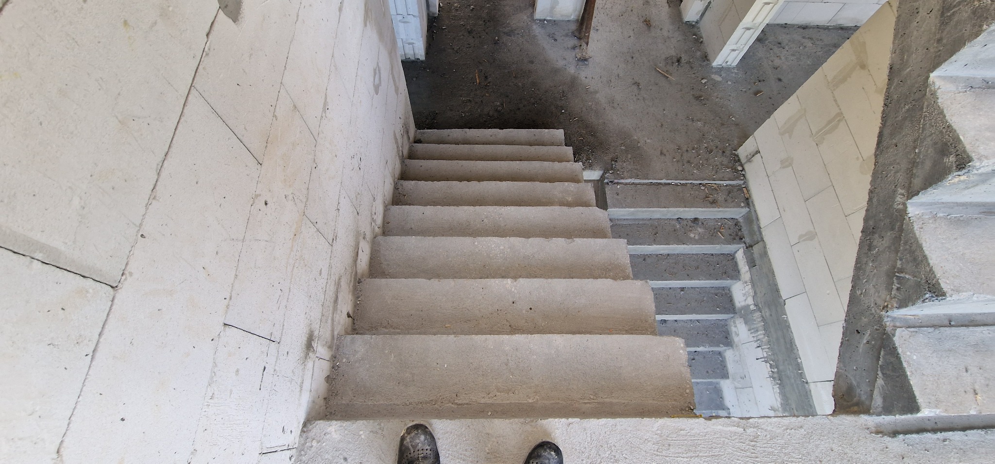 Wnętrze w trakcie budowy: surowe betonowe schody z widocznymi blokami z betonu komórkowego i fragmentem obuwia w dolnej części kadru. Widok z góry.