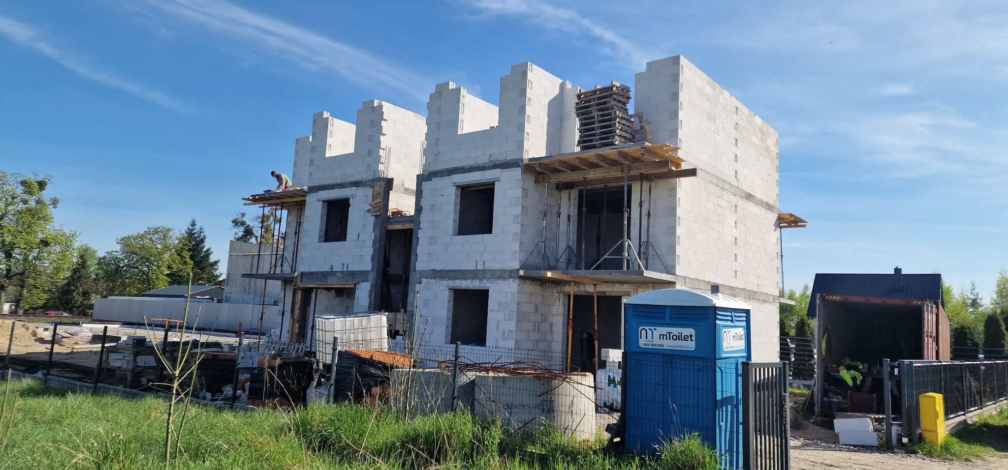 Wznoszenie dwukondygnacyjnego domu z białych bloczków, widoczne rusztowania i pracownik na górze. Częściowo wykończone ściany, okna bez szyb. Błękitne niebo w tle.