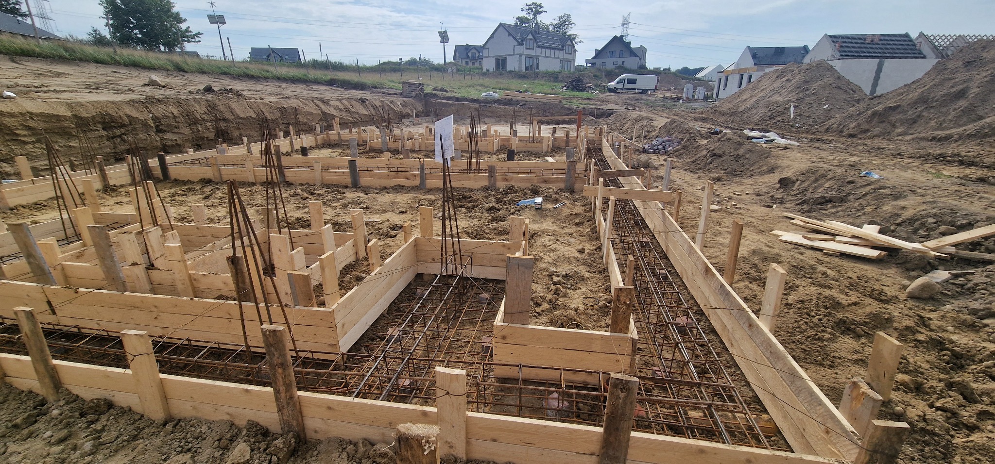 Ziemny wykop z drewnianym szalunkiem i zbrojeniem pod fundamenty domu jednorodzinnego. W tle zabudowa mieszkalna i hałdy ziemi.
