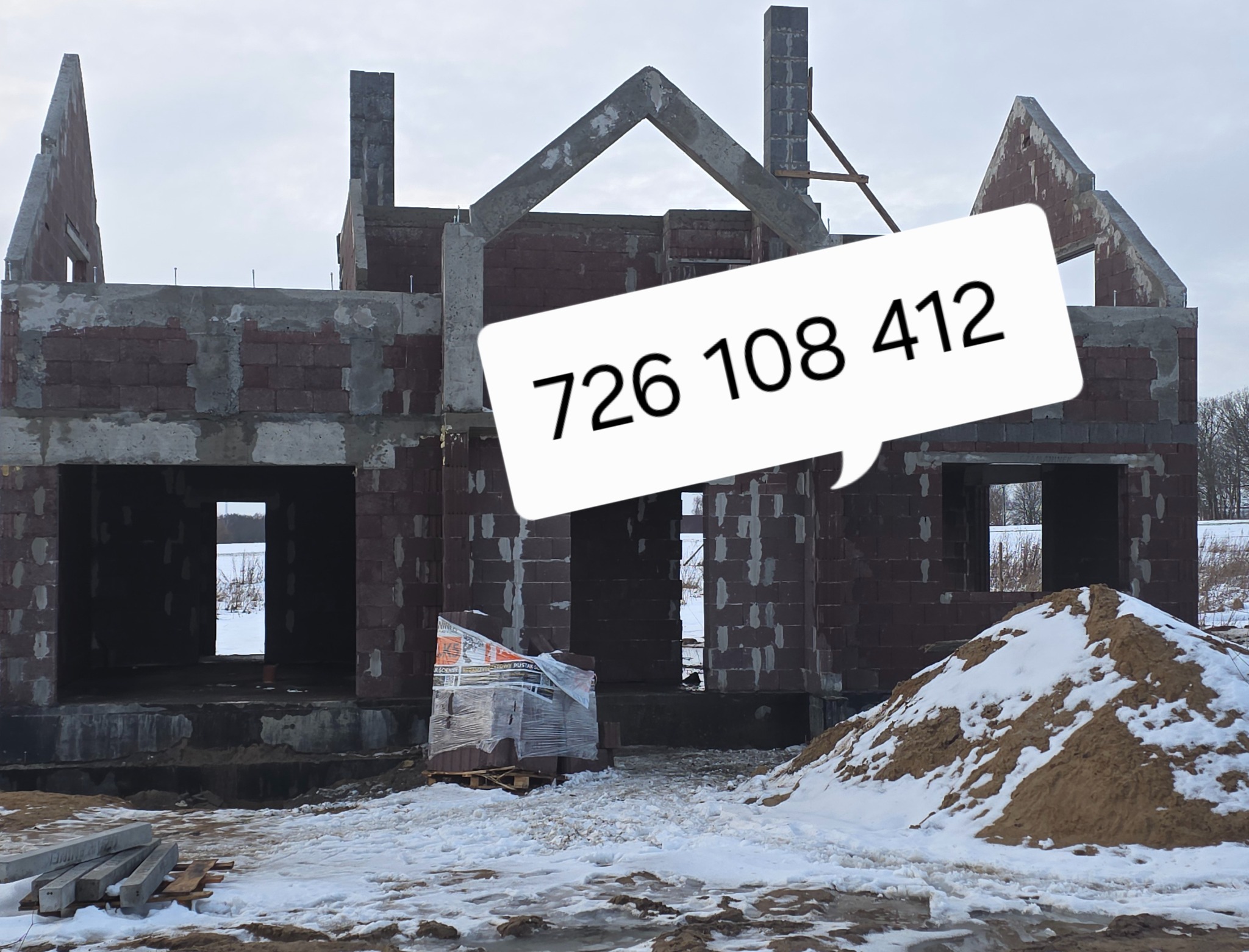 Surowy stan domu z czerwonej cegły w zimowej scenerii, z numerem telefonu w dymku. Widoczne fundamenty, ściany i elementy konstrukcyjne dachu.
