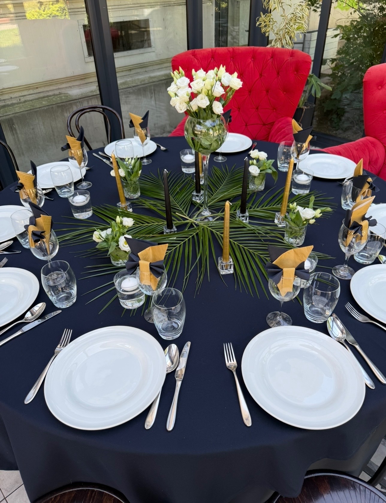 Elegancko nakryty okrągły stół z czarnym obrusem, białymi talerzami, świecami i kwiatami. Dekoracja z liści palmy. Czerwone krzesła w tle.