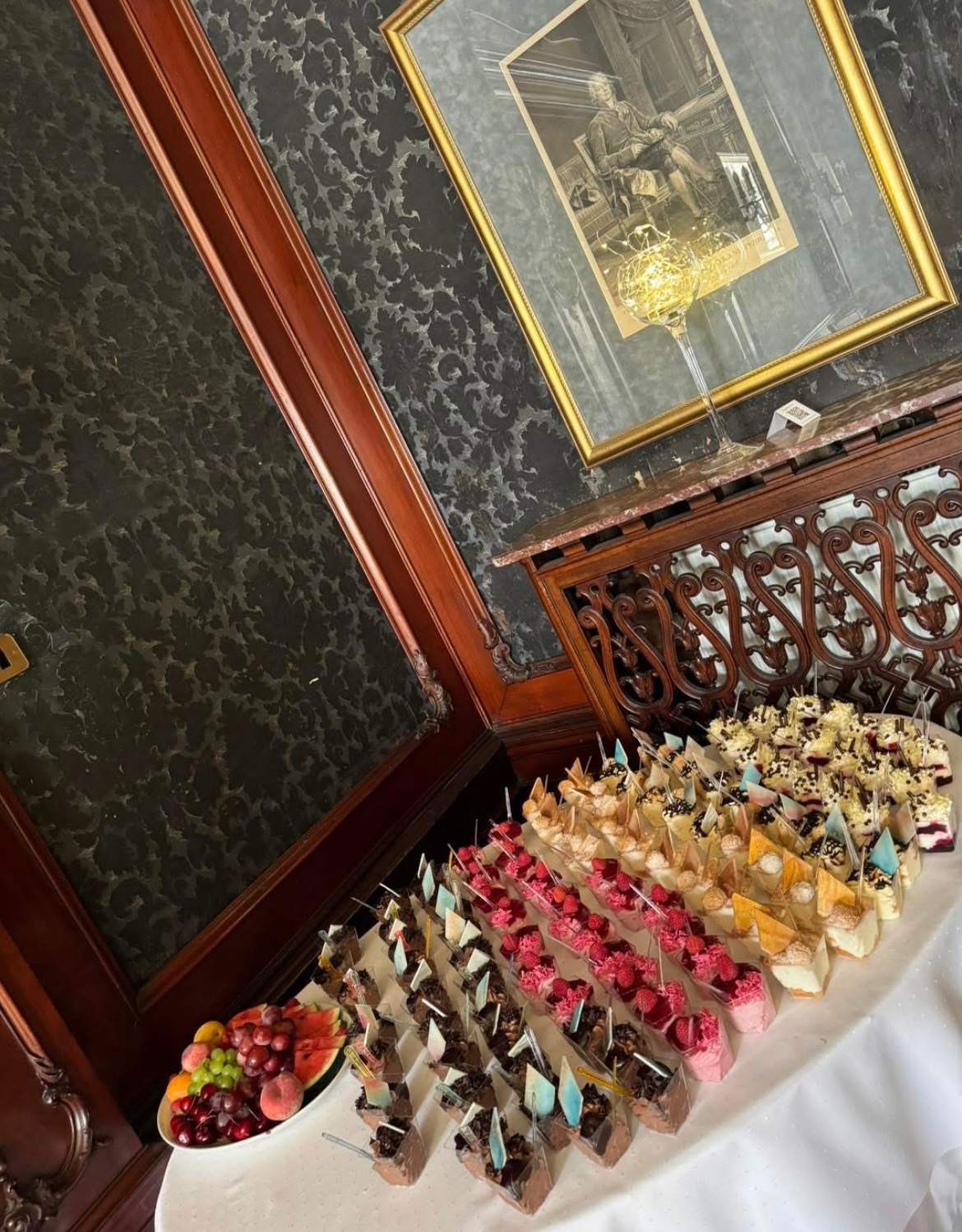 Elegancki stół cateringowy z deserami w szklanych naczyniach i owocami, ustawiony w klasycznym wnętrzu z ciemną tapetą i obrazem w złotej ramie.