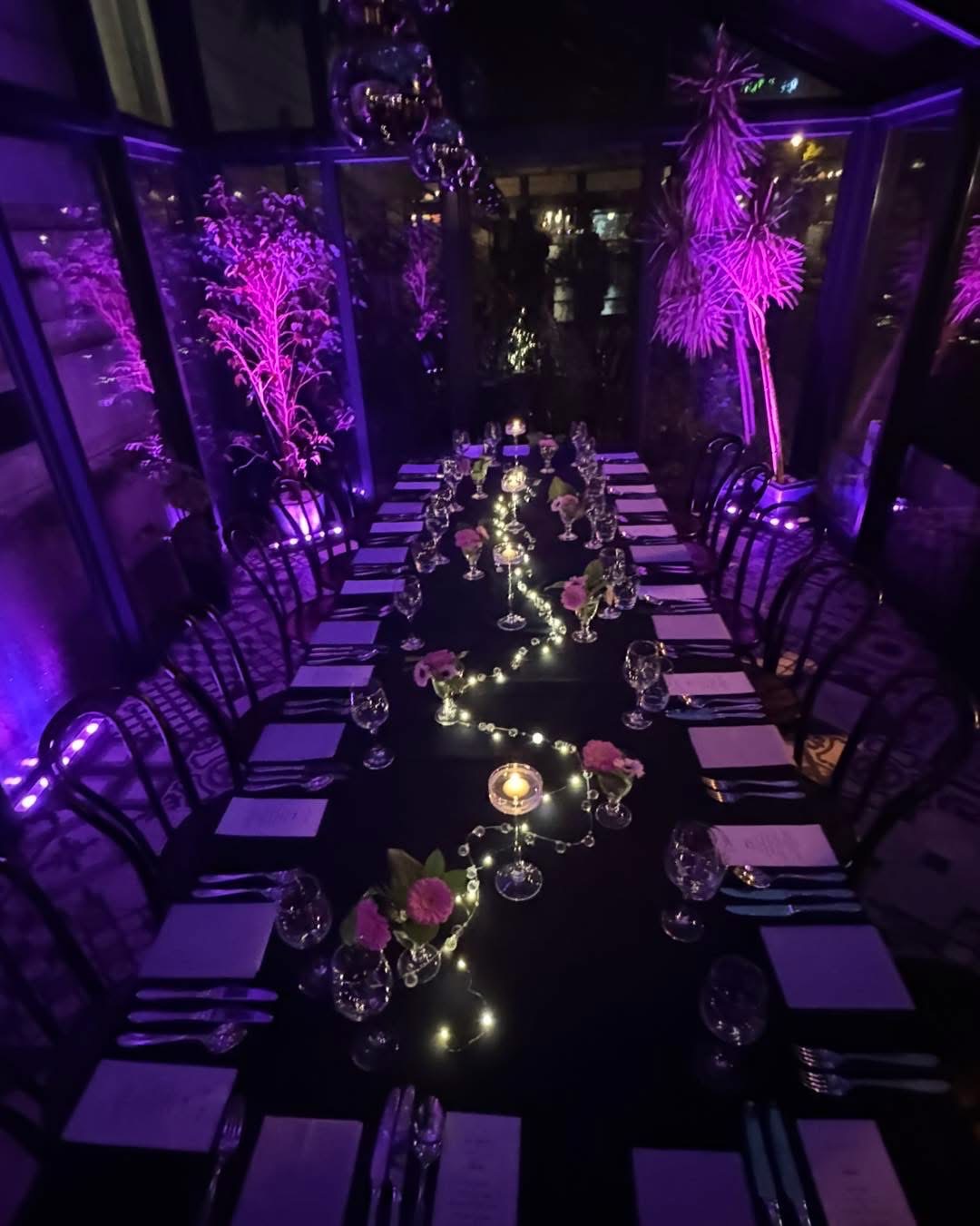 Długi stół z czarnym obrusem, nakryty zastawą i kwiatami, oświetlony fioletowym światłem i girlandami. Dekoracje w szklarni, lustrzana kula na suficie.