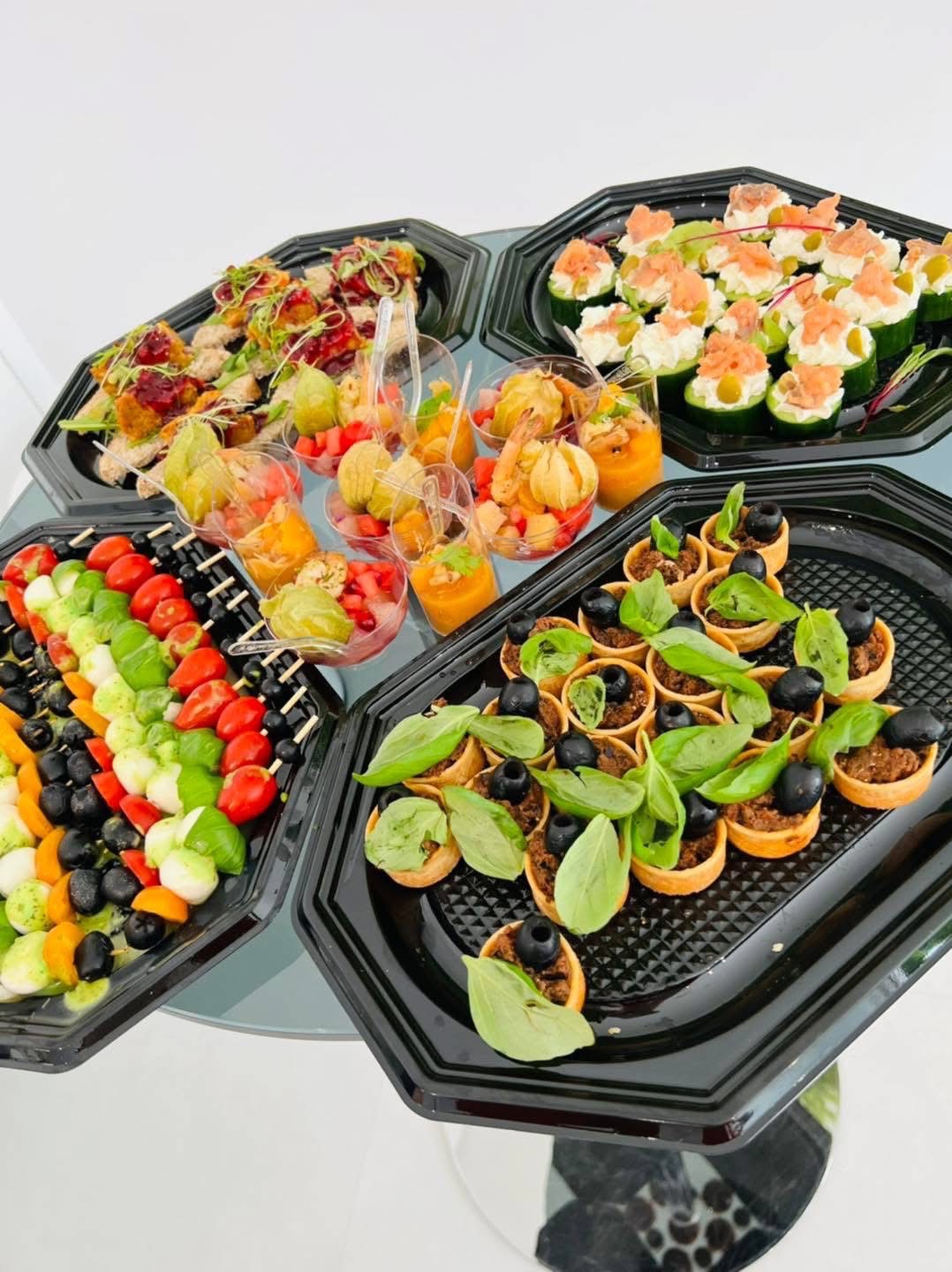 Eleganckie przekąski na czarnych tacach: koreczki z mozzarelli i pomidorów, mini tartaletki z oliwkami, sałatki w szklankach i roladki z łososiem na ogórku.