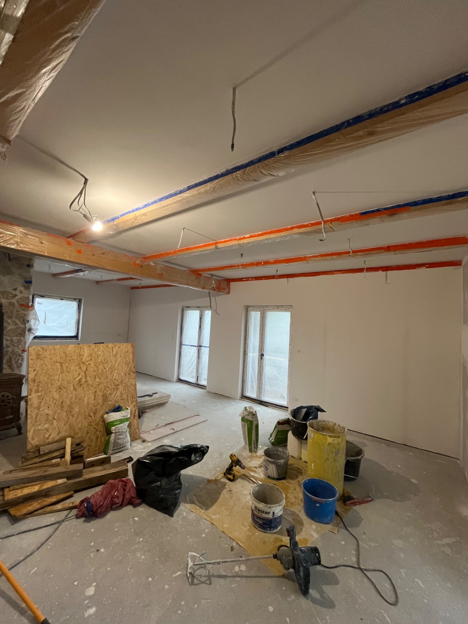 Wnętrze w trakcie prac wykończeniowych: zabezpieczone folią belki, świeżo pomalowane ściany, narzędzia i materiały budowlane na podłodze. Widoczne okna i drzwi.