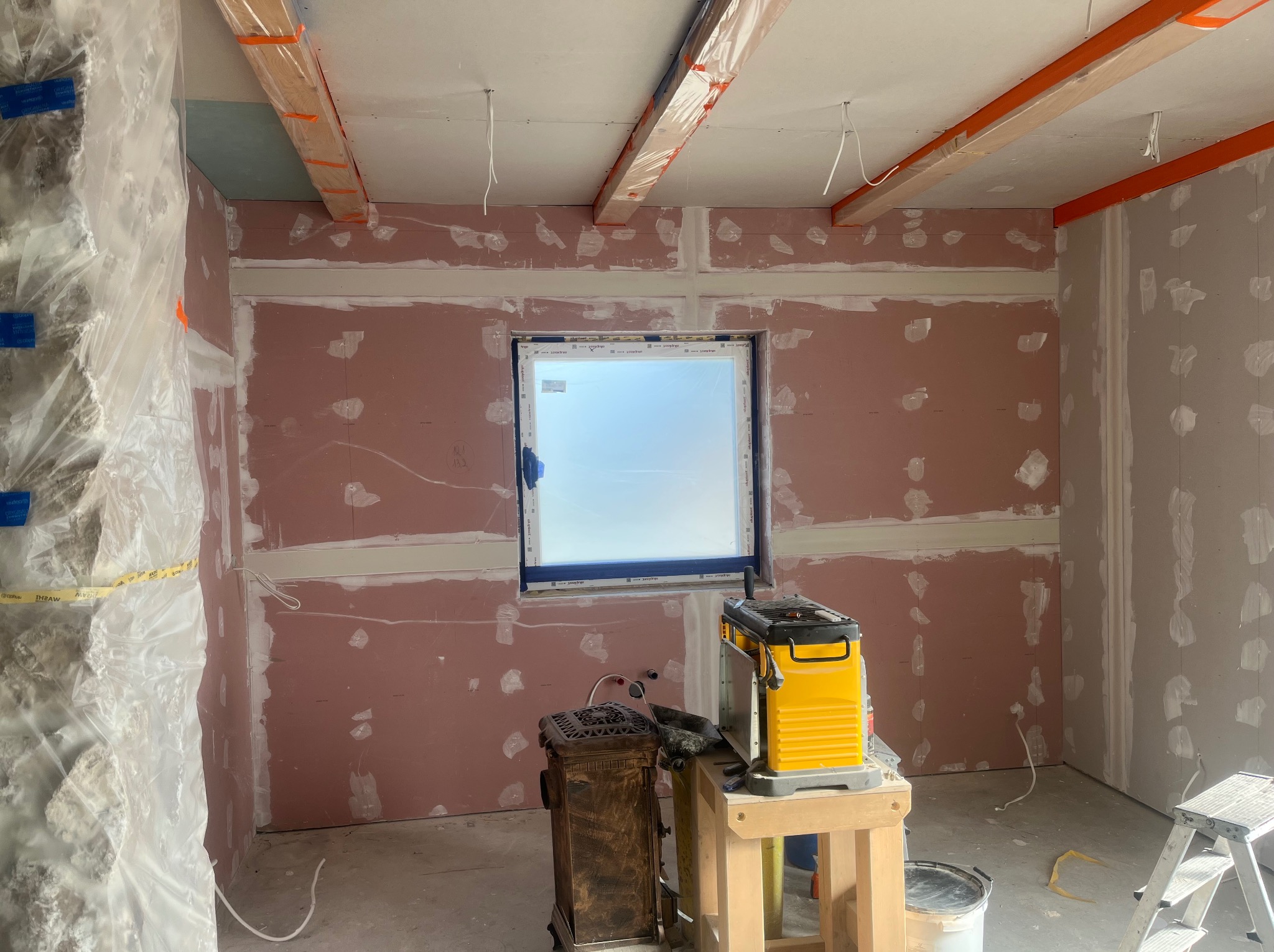 Pokój w trakcie prac wykończeniowych: ściany z płyt gipsowych, widoczne łączenia, okno, narzędzia. Przygotowanie do dalszych prac, np. szpachlowania.