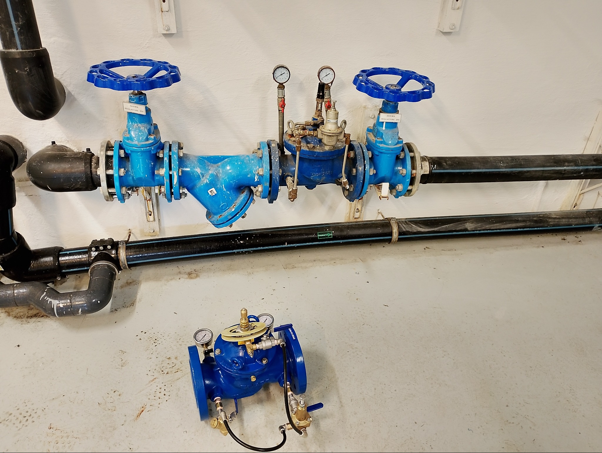 Błękitne zawory z manometrami zamontowane na rurach w przepompowni wody bytowej. Dodatkowy zawór na podłodze. Widoczne elementy instalacji.