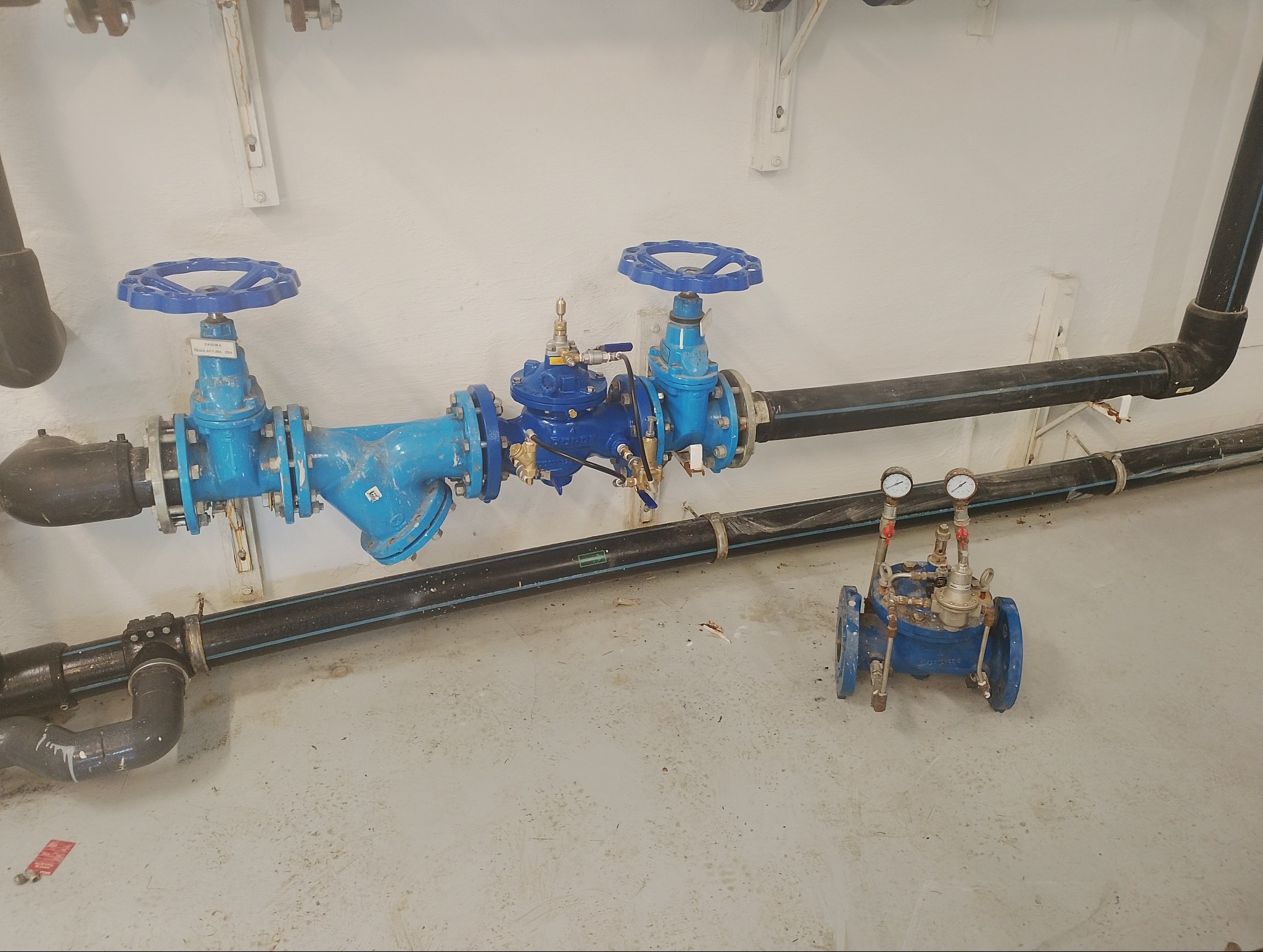 Niebieskie zawory i rury wodociągowe z manometrami, zamontowane na ścianie w przepompowni. Widoczny nowy zawór przed montażem. Instalacja po wymianie zaworu pierwszeństwa.