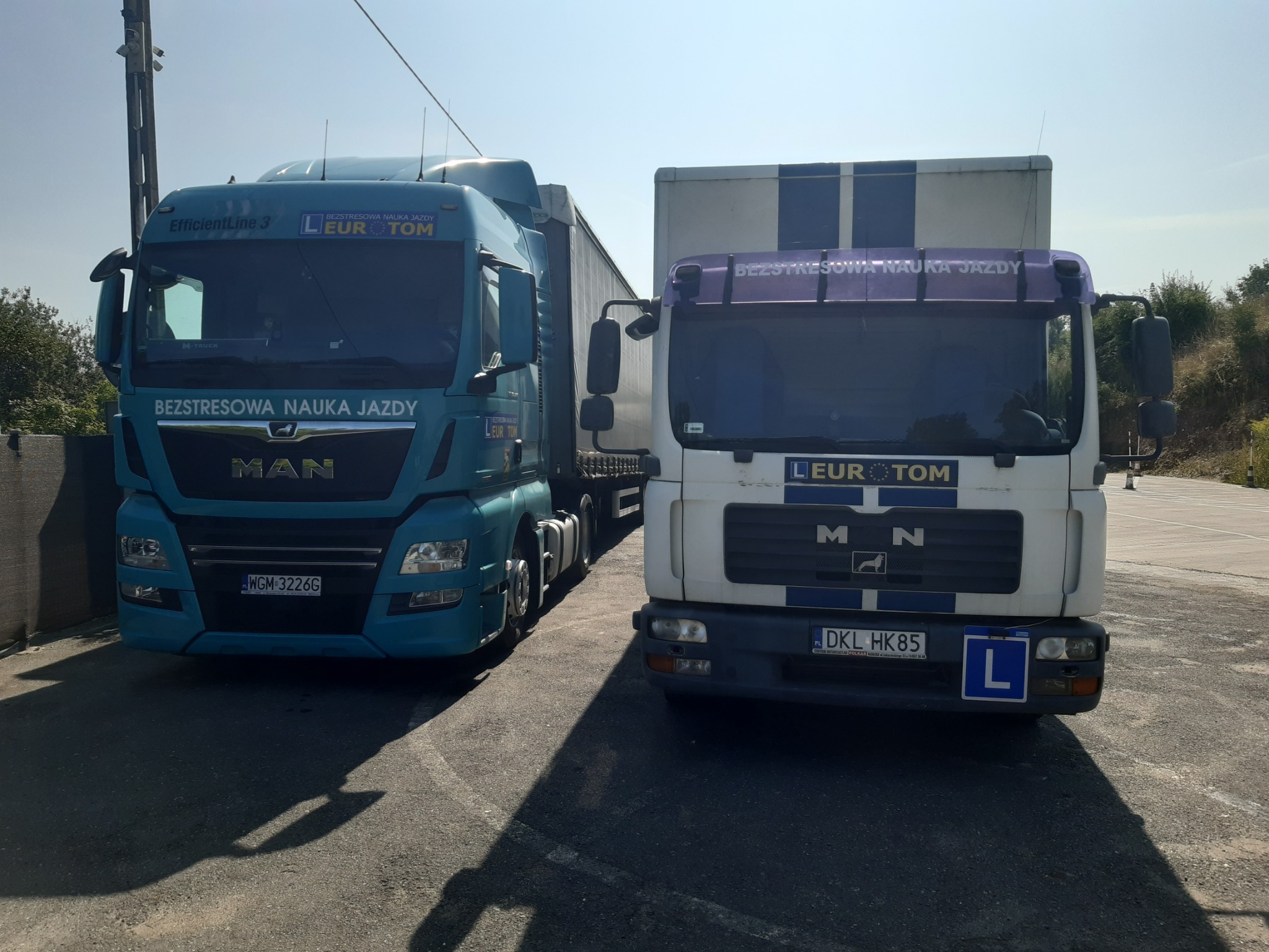 Dwa samochody ciężarowe MAN z oznaczeniami szkoły jazdy 'Bezstresowa Nauka Jazdy' i logo 'EuroTom'. Jeden turkusowy, drugi biały z niebieskimi pasami.