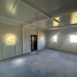 Domtur Garage Garaże z płyt warstwowych - Wnętrze modułowego pomieszczenia z trzema oknami i czarnymi drzwiami. Widoczne białe ściany, sufit z paneli i szara betonowa podłoga. Jasne światło wpada przez okna.