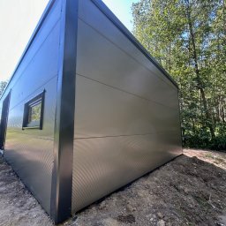 Domtur Garage Garaże z płyt warstwowych - Nowoczesny, szary dom modułowy z oknem, wykonany z paneli, na tle zieleni drzew. Ujęcie z dołu podkreśla bryłę budynku.