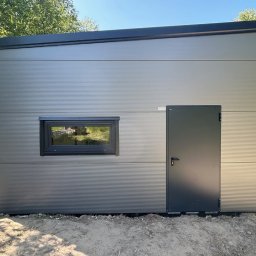Domtur Garage Garaże z płyt warstwowych - Nowoczesny, szary domek modułowy z oknem i drzwiami, wykończony panelami z delikatnym żłobkowaniem, na tle zieleni. Minimalistyczny design.