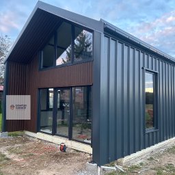 Domtur Garage Garaże z płyt warstwowych - Nowoczesny domek modułowy z szarej blachy trapezowej i brązowymi lamelami drewnopodobnymi na froncie. Duże okna z czarnymi ramami. Widok z zewnątrz, w tle zieleń.