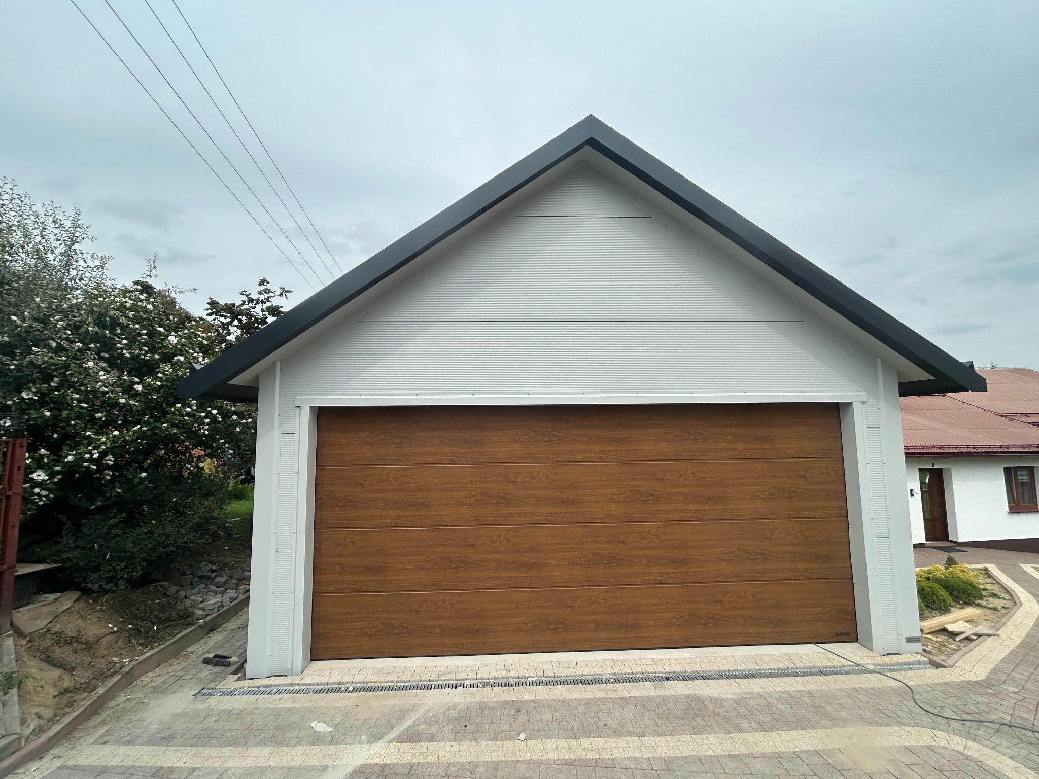 Nowoczesny garaż blaszany z dwuspadowym dachem i bramą segmentową w kolorze drewna. Białe ściany z płyty PIR z ukrytym łącznikiem. Widok z zewnątrz, kostka brukowa.