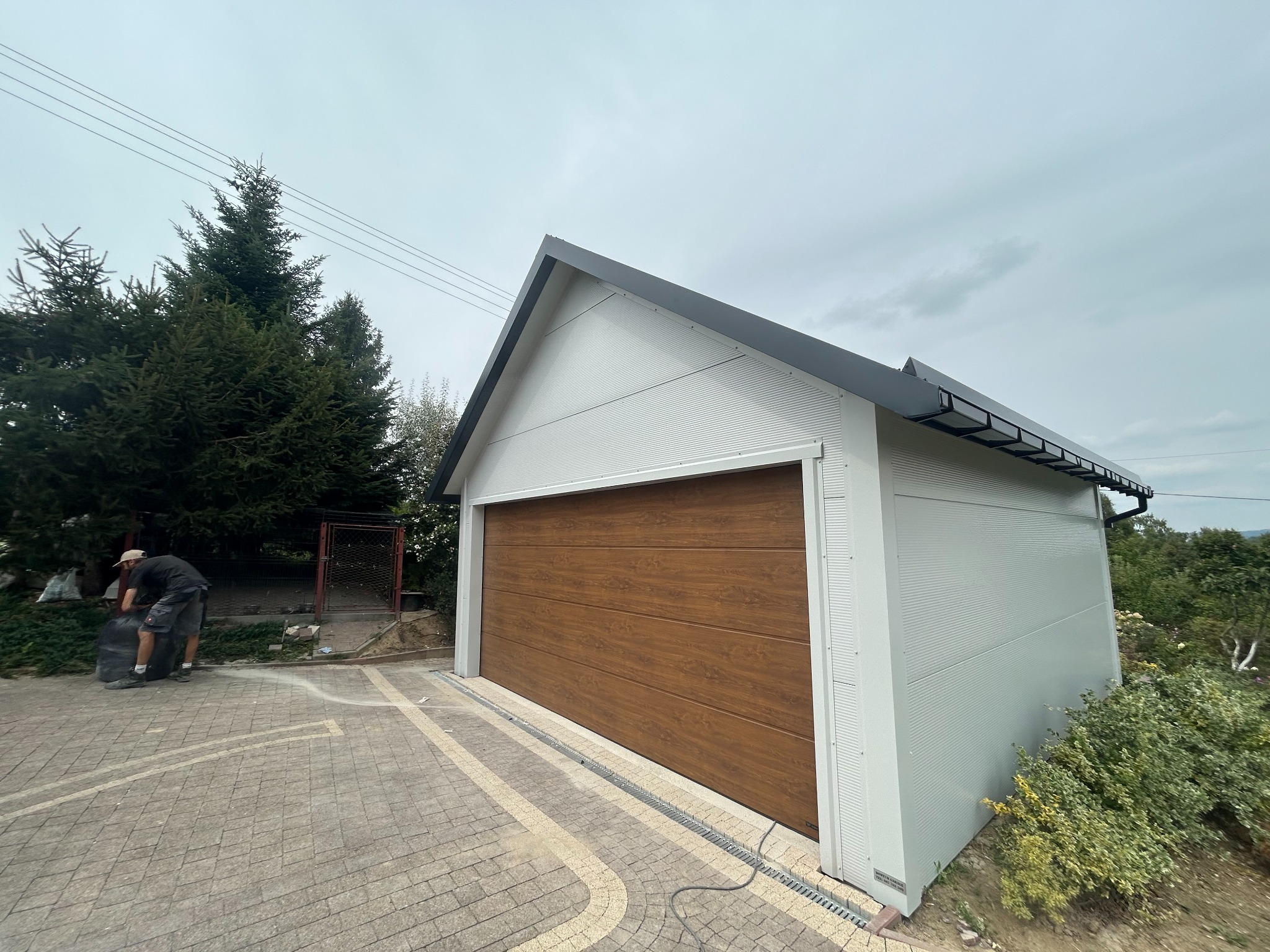 Nowoczesny garaż blaszany z brązową bramą segmentową, widok z zewnątrz. Osoba sprząta teren obok. Szara elewacja garażu i ciemna dachówka. Kostka brukowa przed garażem.