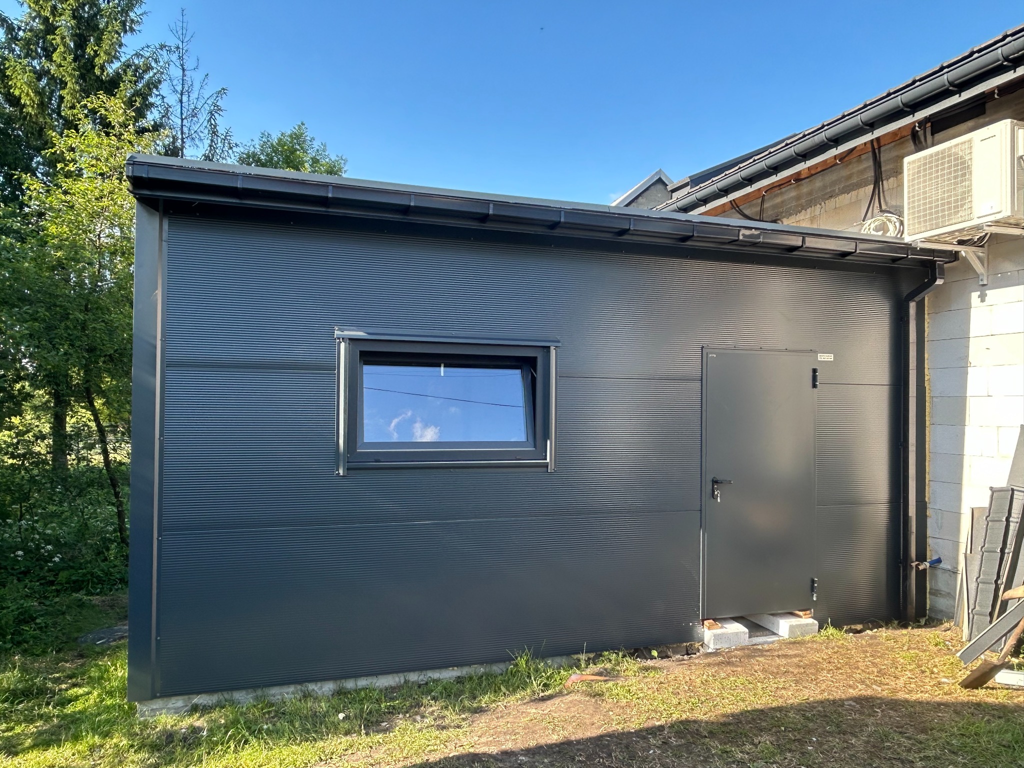 Nowoczesny, antracytowy budynek z płyty warstwowej z oknem i drzwiami, przylegający do budynku mieszkalnego. Widoczne elementy instalacji klimatyzacji.