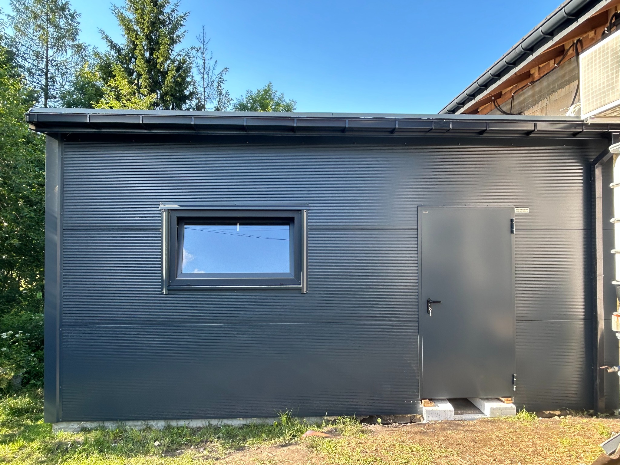 Nowoczesny, grafitowy garaż blaszany z oknem i drzwiami, umieszczony na betonowych bloczkach, otoczony zielenią i błękitnym niebem.