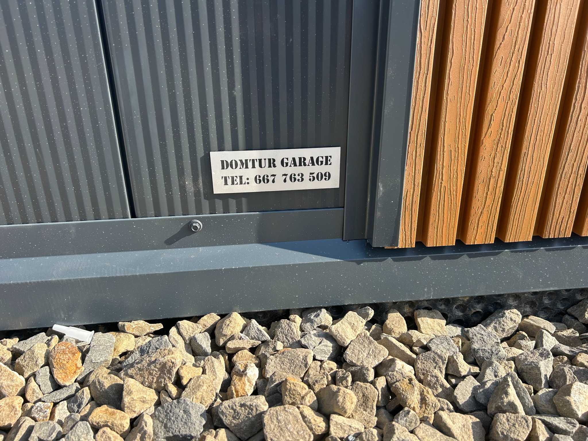 Szczegółowe ujęcie garażu blaszanego z elementami imitującymi drewno i tabliczką z nazwą firmy oraz numerem telefonu, otoczonego kamieniami.