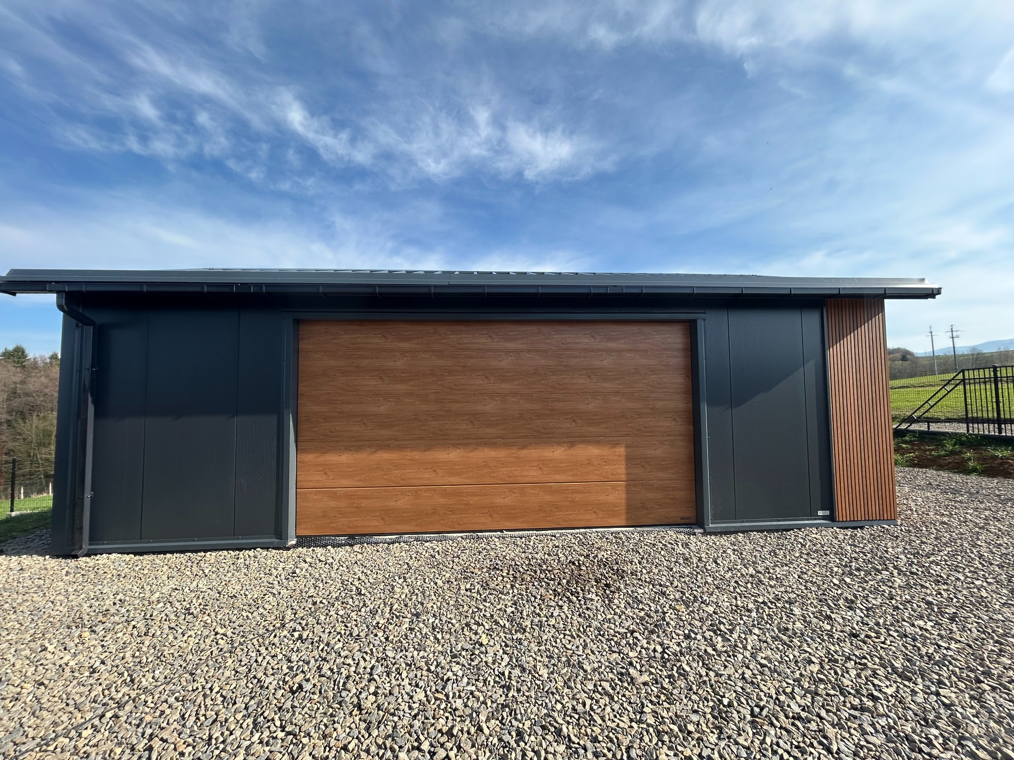 Nowoczesny garaż blaszany z bramą segmentową w kolorze drewna i grafitowymi ścianami, otoczony żwirem, na tle błękitnego nieba z chmurami.