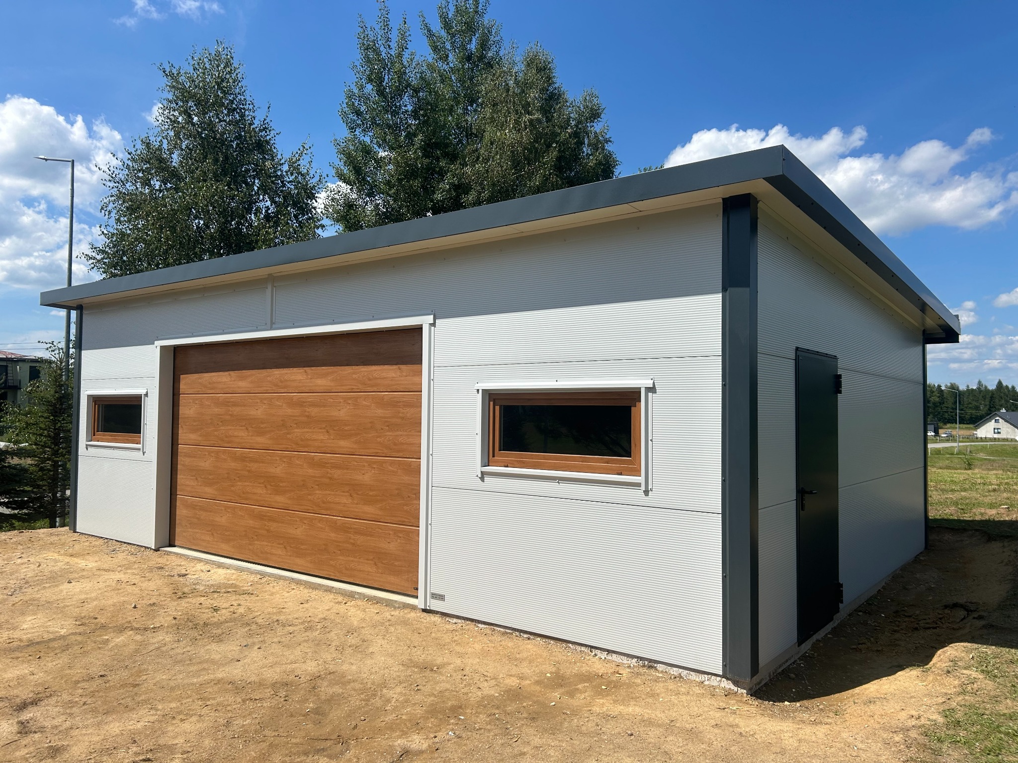 Nowoczesny garaż blaszany z bramą segmentową w kolorze drewna i oknami, wykonany z płyty warstwowej, na tle błękitnego nieba z chmurami.