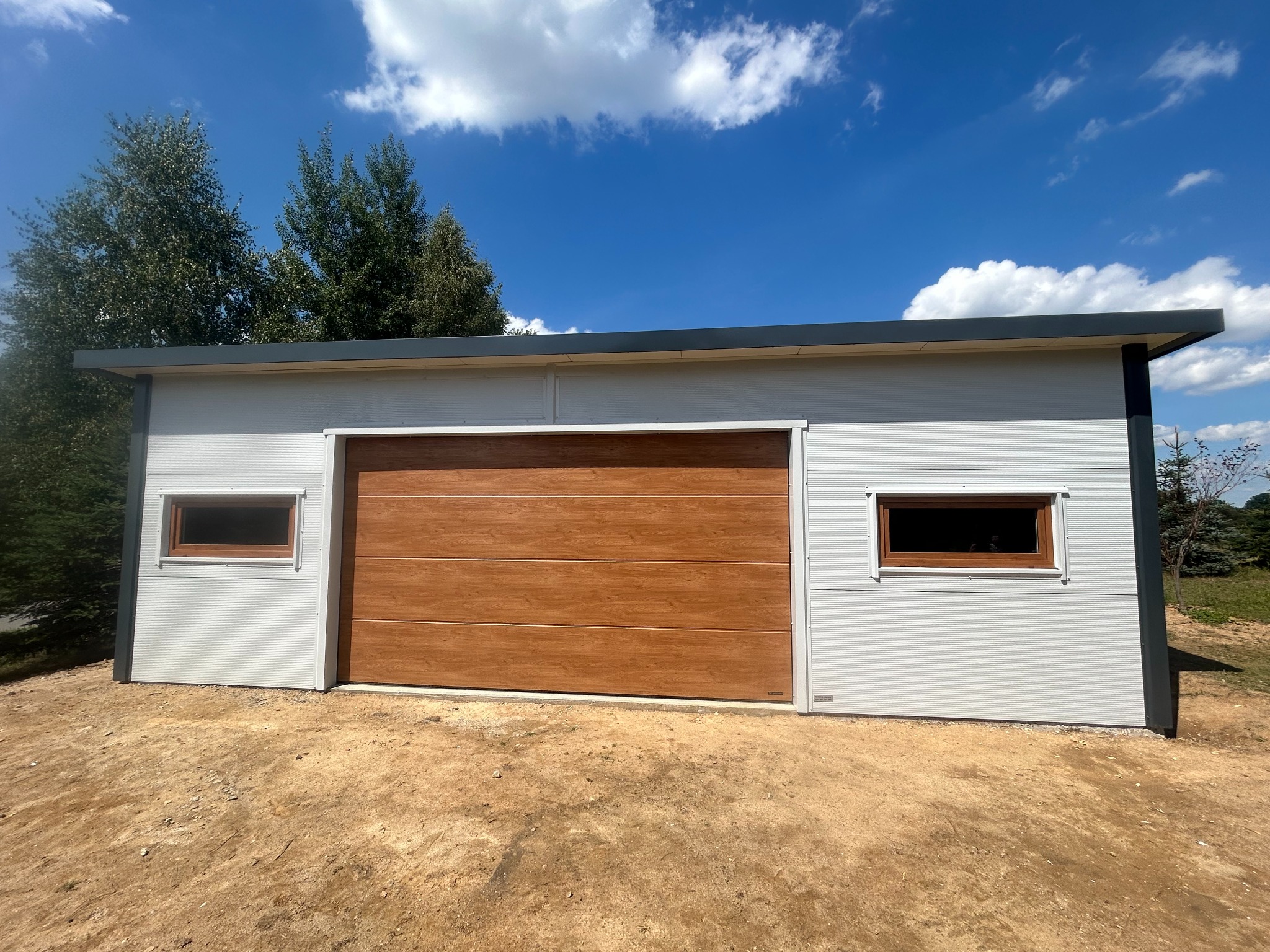 Nowoczesny garaż blaszany z bramą segmentową w kolorze drewna i oknami, na tle błękitnego nieba z chmurami. Widoczne drzewa i ziemiste podłoże.