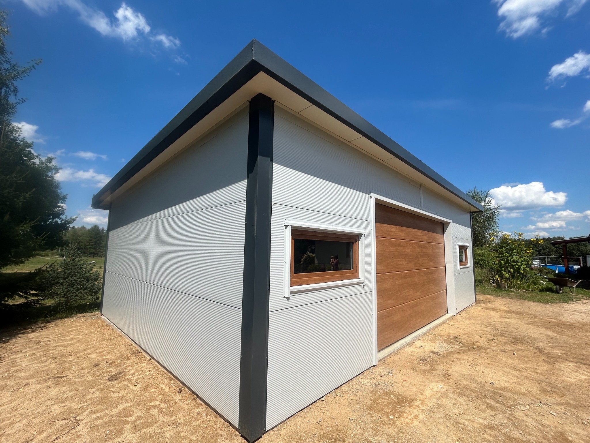 Nowoczesny garaż blaszany z panelową bramą w odcieniu drewna, oświetlony jasnym słońcem na tle błękitnego nieba z lekkimi obłokami. Widok z perspektywy narożnika.