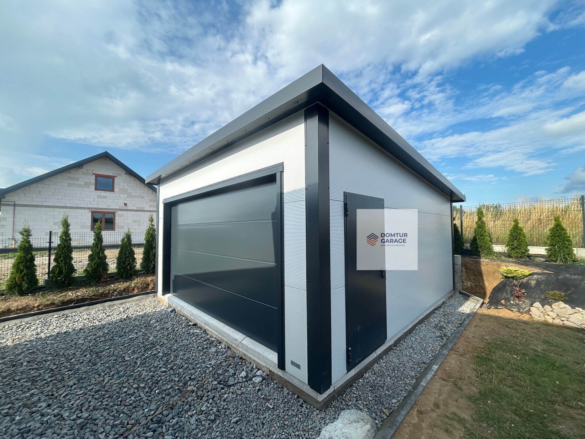Nowoczesny garaż blaszany z grafitową bramą segmentową i drzwiami, obok domu jednorodzinnego, otoczony zielenią i kamiennym podłożem. Widok z boku.