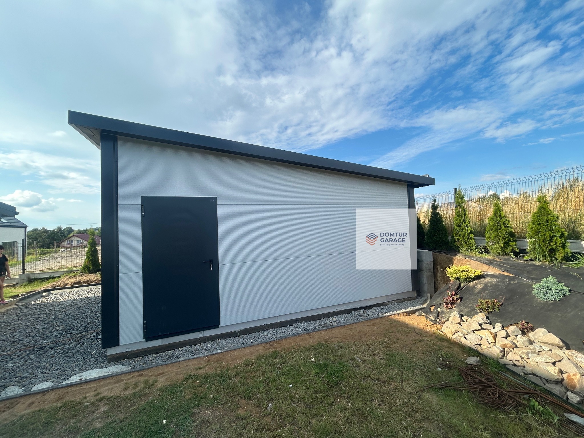 Nowoczesny garaż blaszany z jednospadowym dachem i czarnymi detalami, otoczony zielenią i kamieniami. Widok z boku, podkreślający minimalistyczny design i estetyczne wkomponowanie w otoczenie.